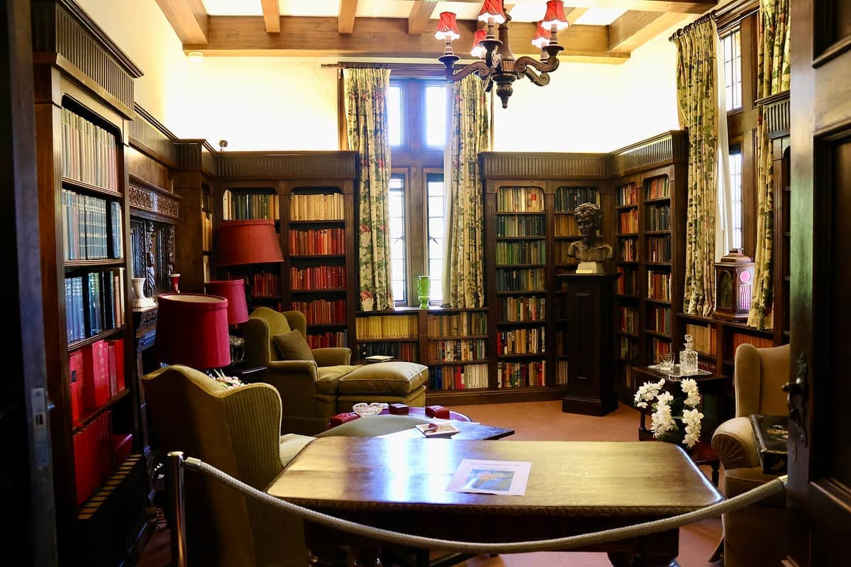 Biblioteca con paneles de madera en Carrick Hill con estantes llenos de libros, ventana grande, cortinas florales, sillones verdes y una araña de luces que proporciona luz cálida.