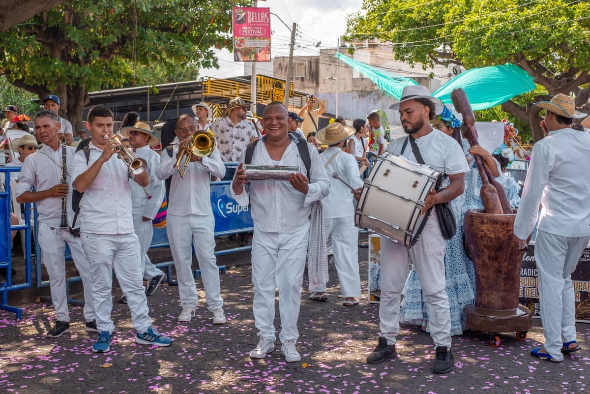 A group of musicians and dancers in white clothing performing with drums and brass instruments outdoors during a festive street celebration.