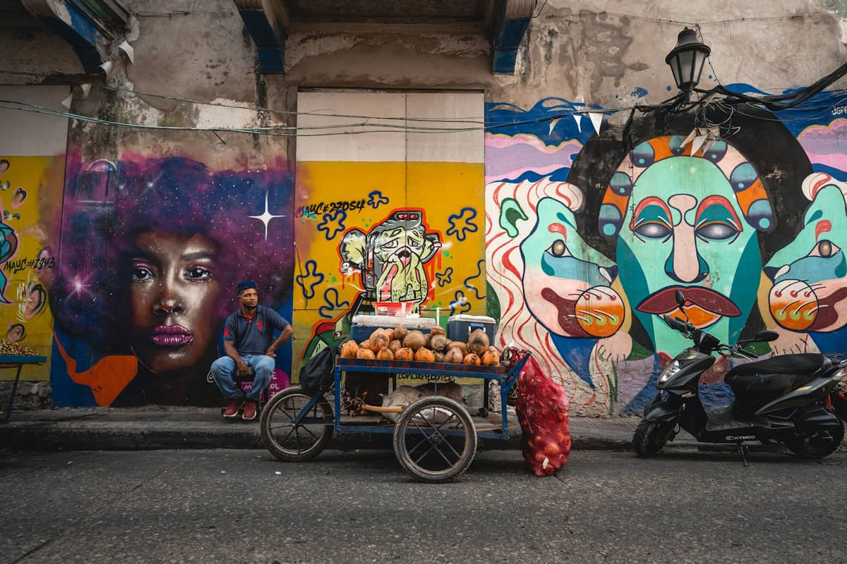 Street vendor with coconut cart in front of colorful Afro-Colombian murals in Cartagena, highlighting local food culture and heritage.