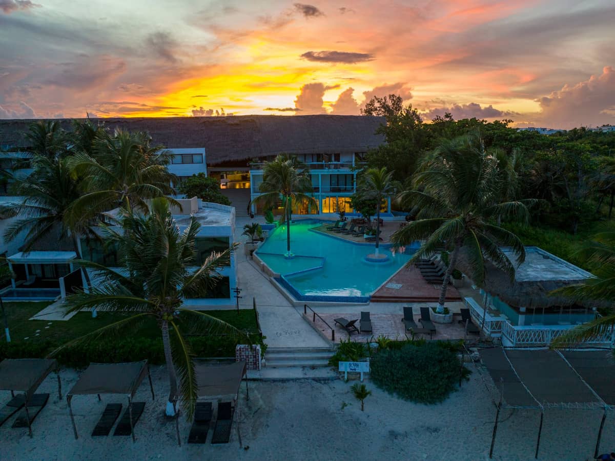 Aerial view of a beachfront resort complex with pools, palm trees, and a main building at sunset, evoking tropical all-inclusive vibes.