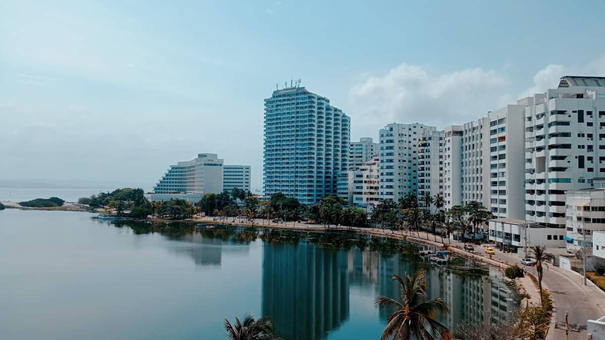 Modern beachfront resort hotels in Bocagrande, Cartagena, reflected in a calm lagoon with palm trees and blue sky.