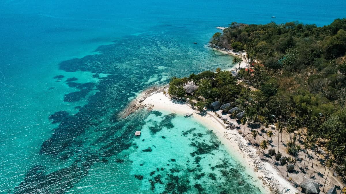 Aerial view of a white sand beach on a lush green island bordered by turquoise water and visible coral reefs.