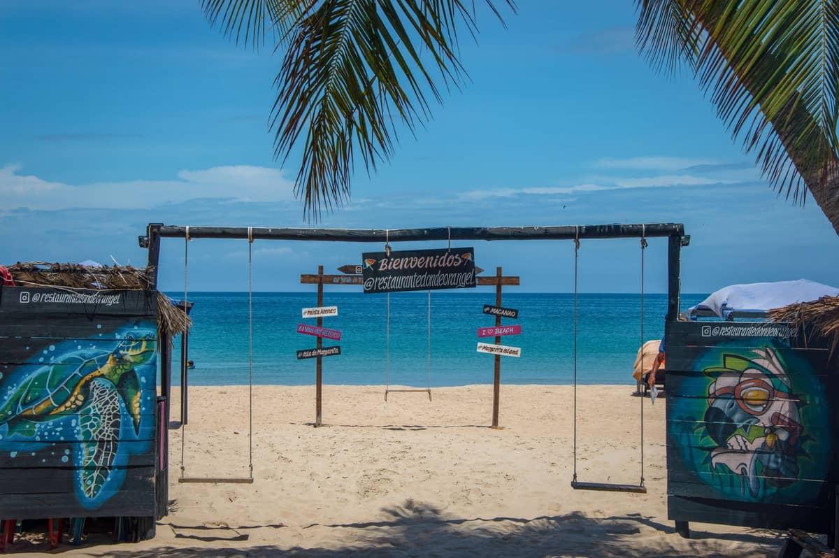 Colorful entrance with swings, artwork, and Spanish welcome sign on a tropical Caribbean beach with turquoise water and palm trees