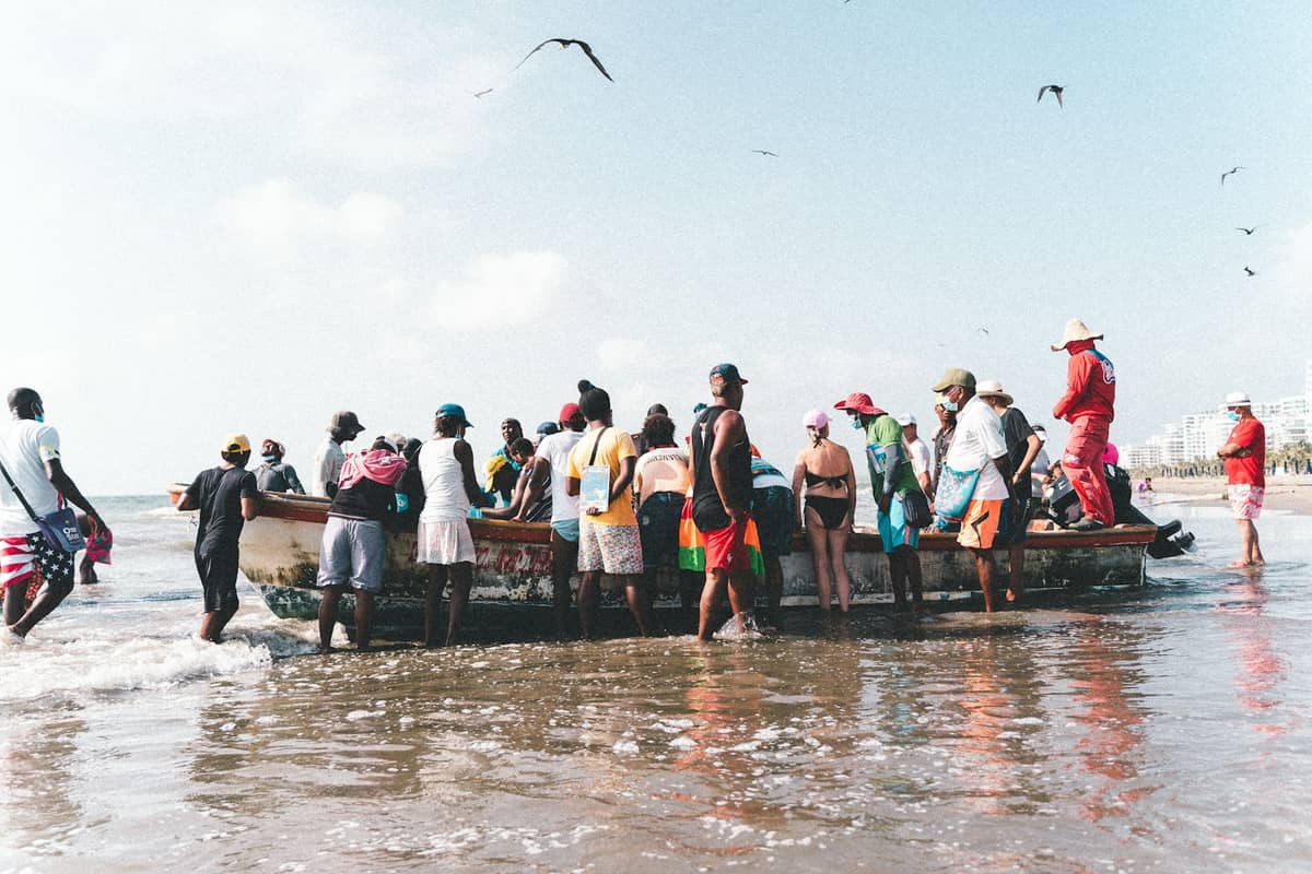 A group of people standing in shallow water around a small fishing boat on a beach, with seabirds flying overhead.