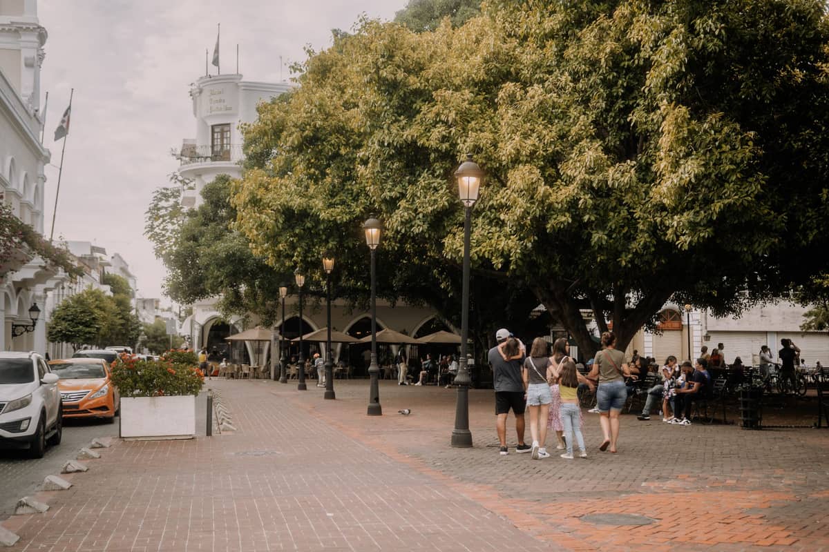 Group of tourists gathering in a historic colonial plaza with white buildings and large trees, possibly preparing for a tour or exploring independently.