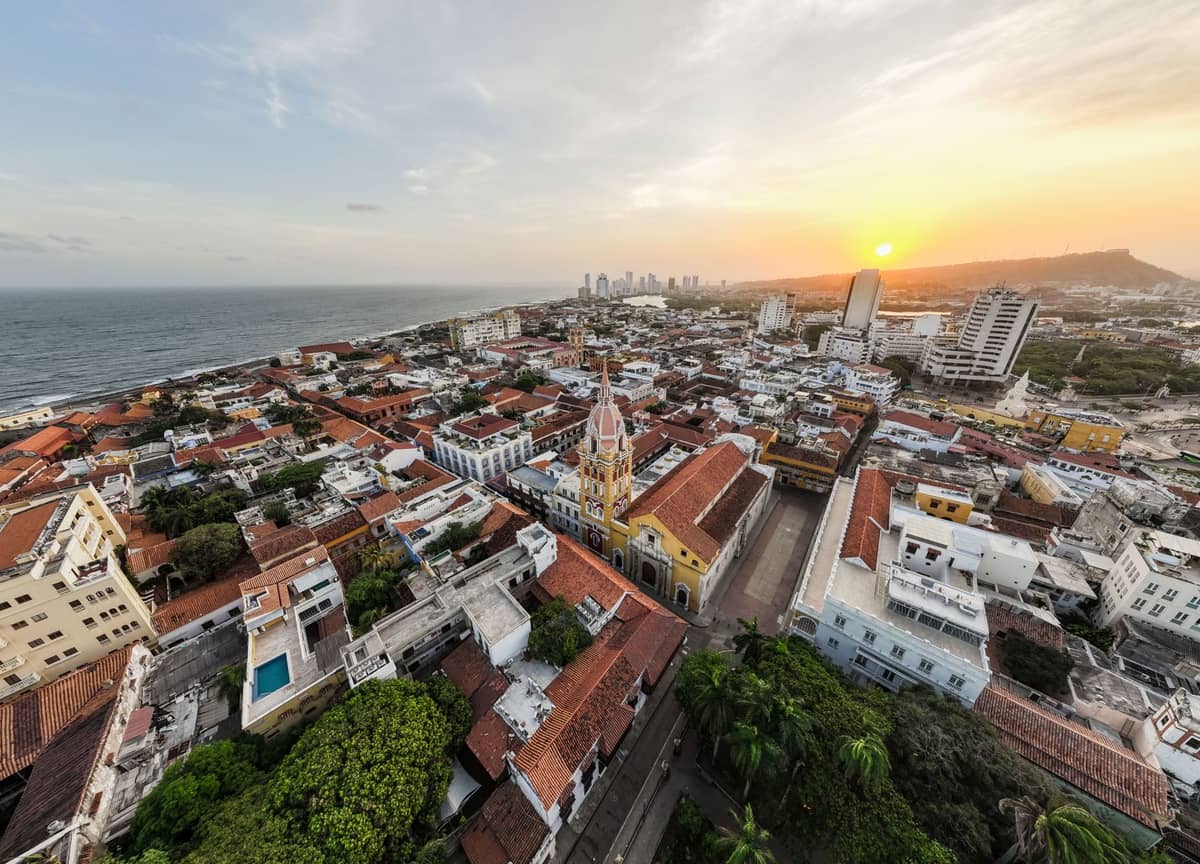 A panoramic aerial view of Cartagena's historic walled city at sunset, with colonial buildings and the Cathedral of Cartagena clearly visible near the center.