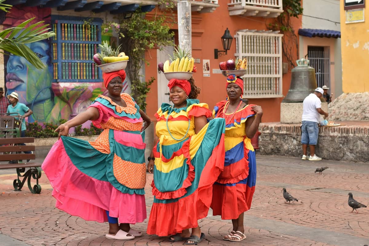Three women in colorful traditional dresses pose in a Cartagena square with colonial buildings and murals in the background.