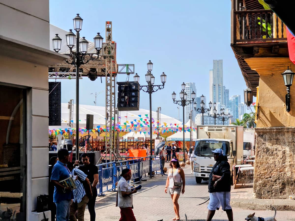 Street scene in Cartagena with people walking, vendors, colonial lampposts, market stalls, and the skyline in the background.
