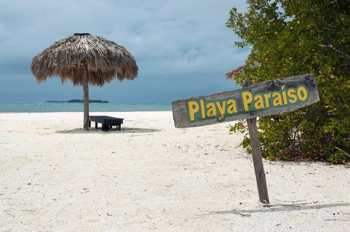 A sign reading 'Playa Paraíso' on a sandy beach with a thatched umbrella, clear turquoise sea, and some greenery in Cartagena's Rosario Islands.