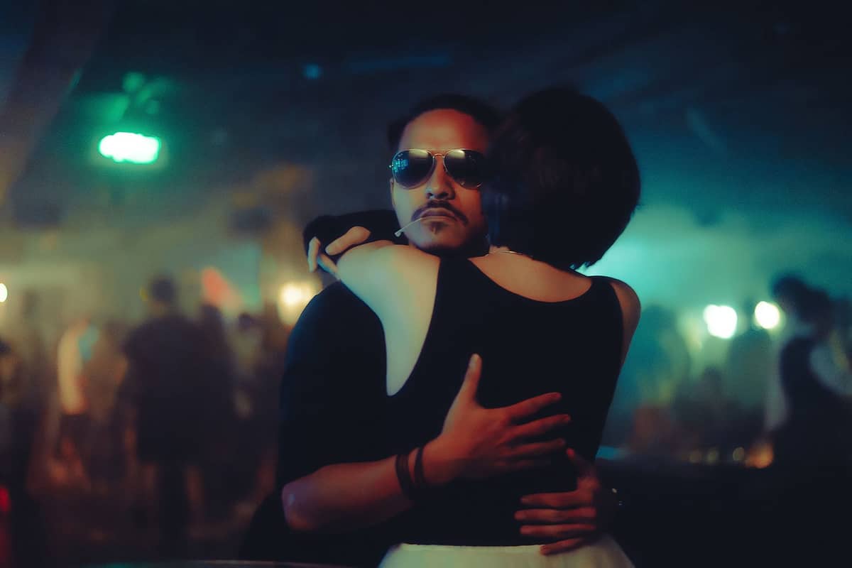 A couple dancing closely in a dimly lit nightclub with other clubgoers and colorful lights in the background.