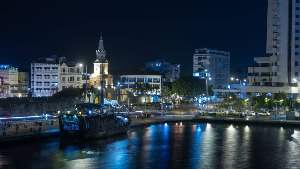 Nighttime view of Cartagena’s waterfront with the historic clock tower, city lights reflecting in the water, and lively night atmosphere.