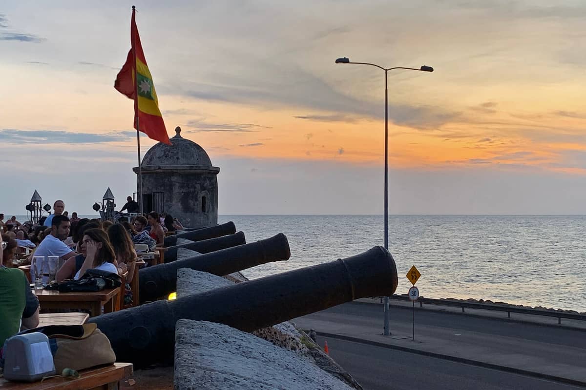People sitting at tables along Cartagena's famous city walls with cannons, sunset over the Caribbean Sea, and a flag in the background.