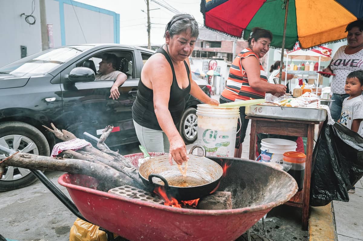 Female street vendor in Cartagena frying fritos over wood fire in a wheelbarrow, surrounded by other locals and colorful market scene.