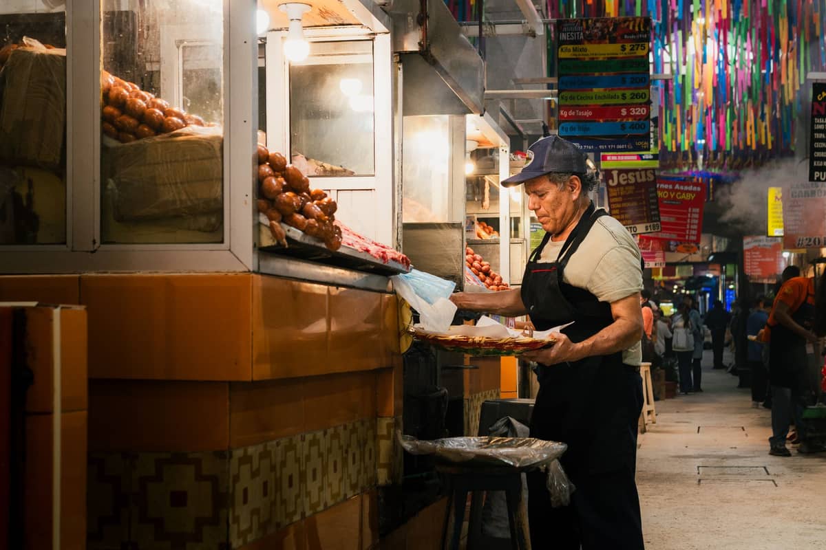 Indoor market scene with a vendor in an apron working at a food stall surrounded by baked goods and festive hanging decorations.