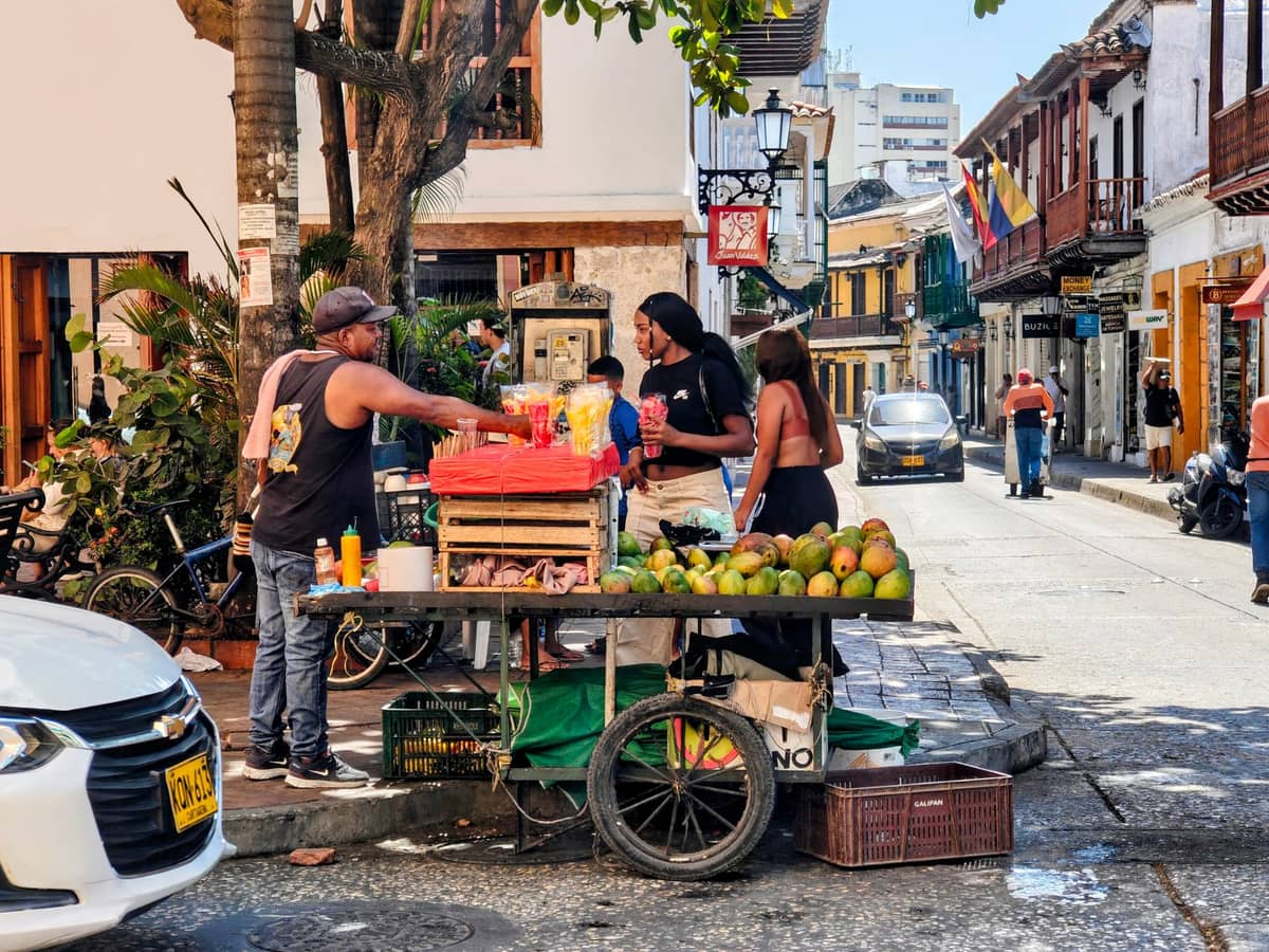 Street food cart in Cartagena de Indias with people buying snacks, tropical fruit, and colorful colonial buildings in the background on a lively city street.
