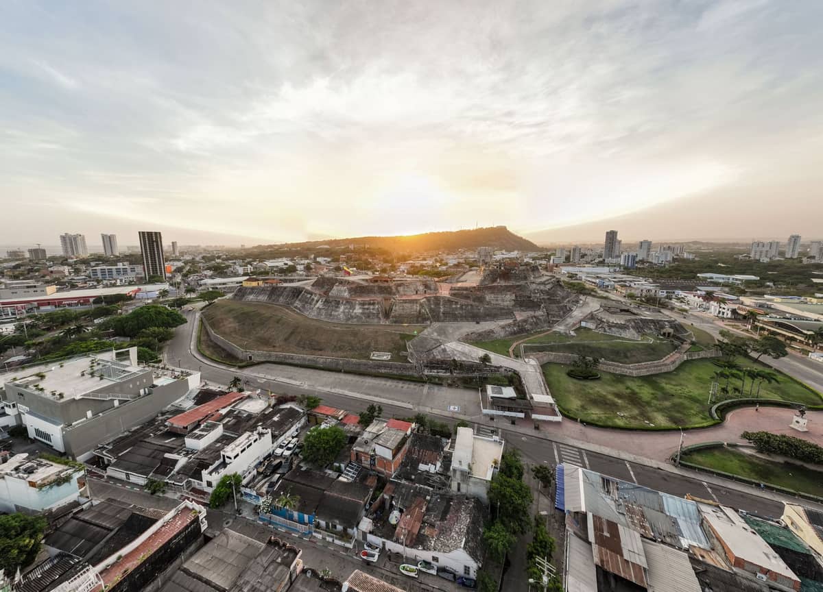 Aerial view of a massive stone fortress with ramparts, surrounded by city buildings and greenery at sunset.