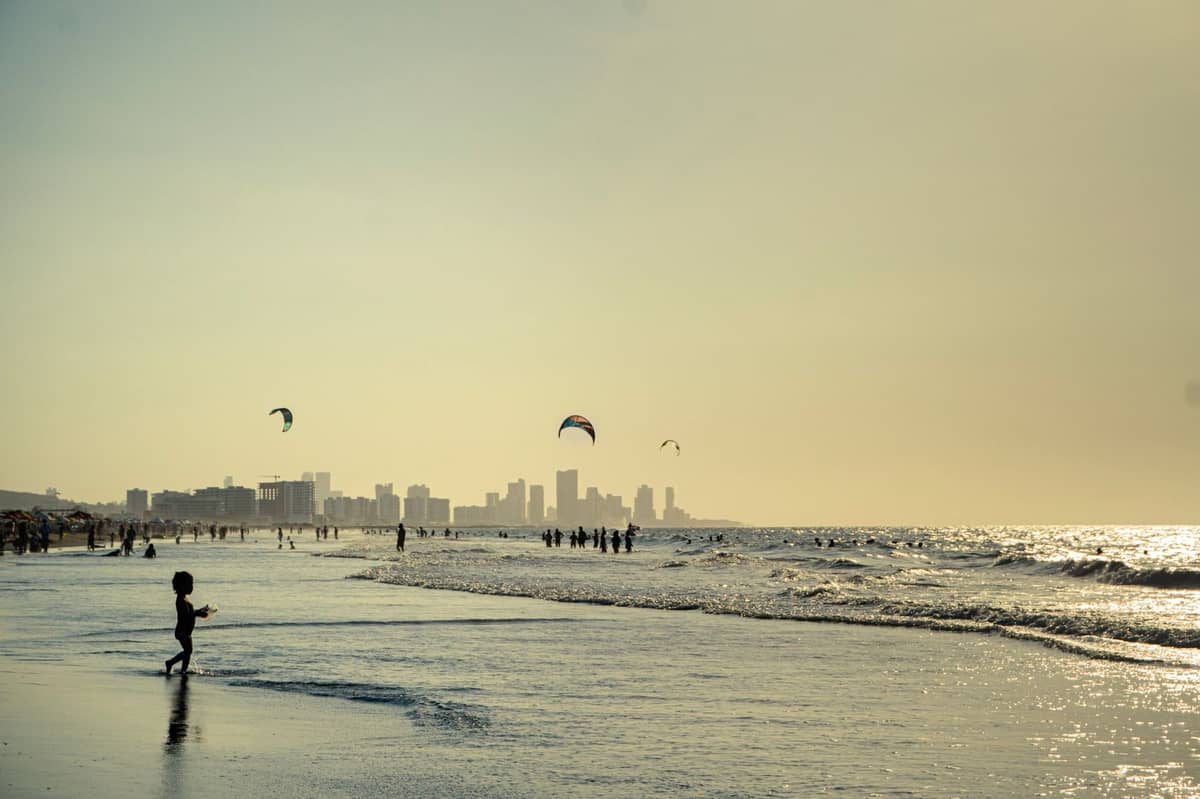Wide sandy beach with people wading and playing in the gentle surf, kites in the sky, Cartagena skyline in the distance.