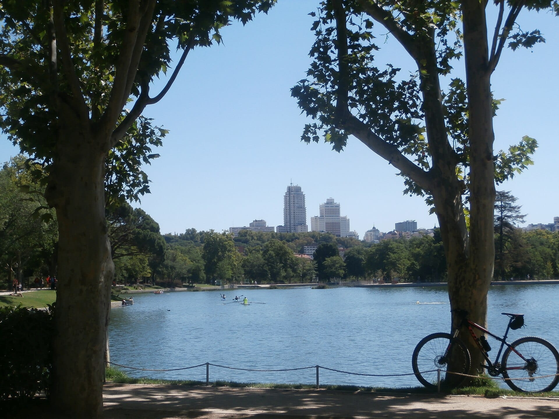 Vista del lago della Casa de Campo a Madrid con alberi rigogliosi, grattacieli della città sullo sfondo e una bicicletta appoggiata sul bordo dell'acqua.