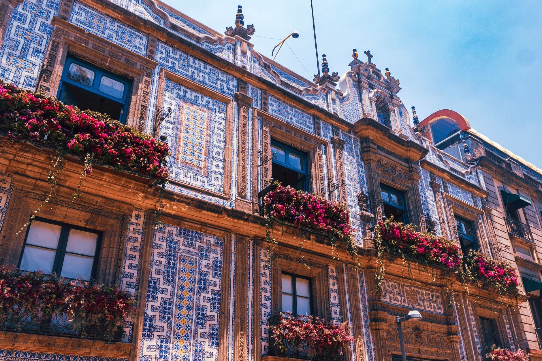 Facciata esterna della Casa de los Azulejos ricoperta di intricate piastrelle Talavera bianche e blu, con fiori in fiore sui balconi delle finestre sotto un cielo luminoso.