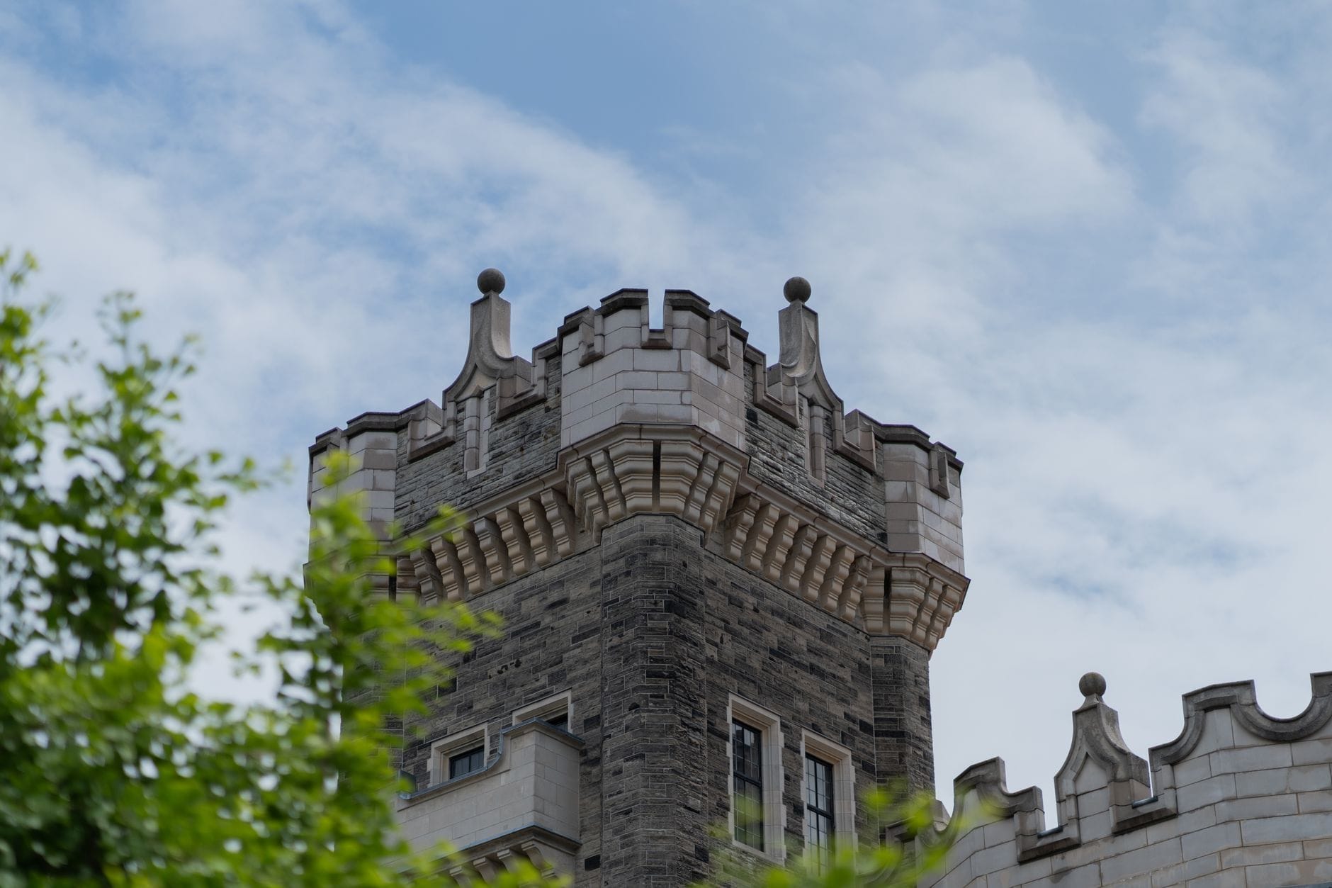 มุมซูมใกล้หอคอยหินเอกลักษณ์ของ Casa Loma รายล้อมด้วยกิ่งไม้เขียวและท้องฟ้ามีเมฆบางๆ