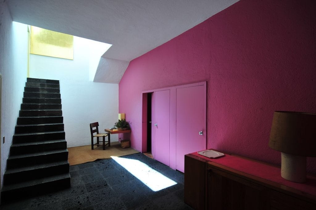 Interno della Casa Luis Barragán con una vivace parete rosa, arredi minimalisti, una scala scura e luce naturale che filtra dall'alto.