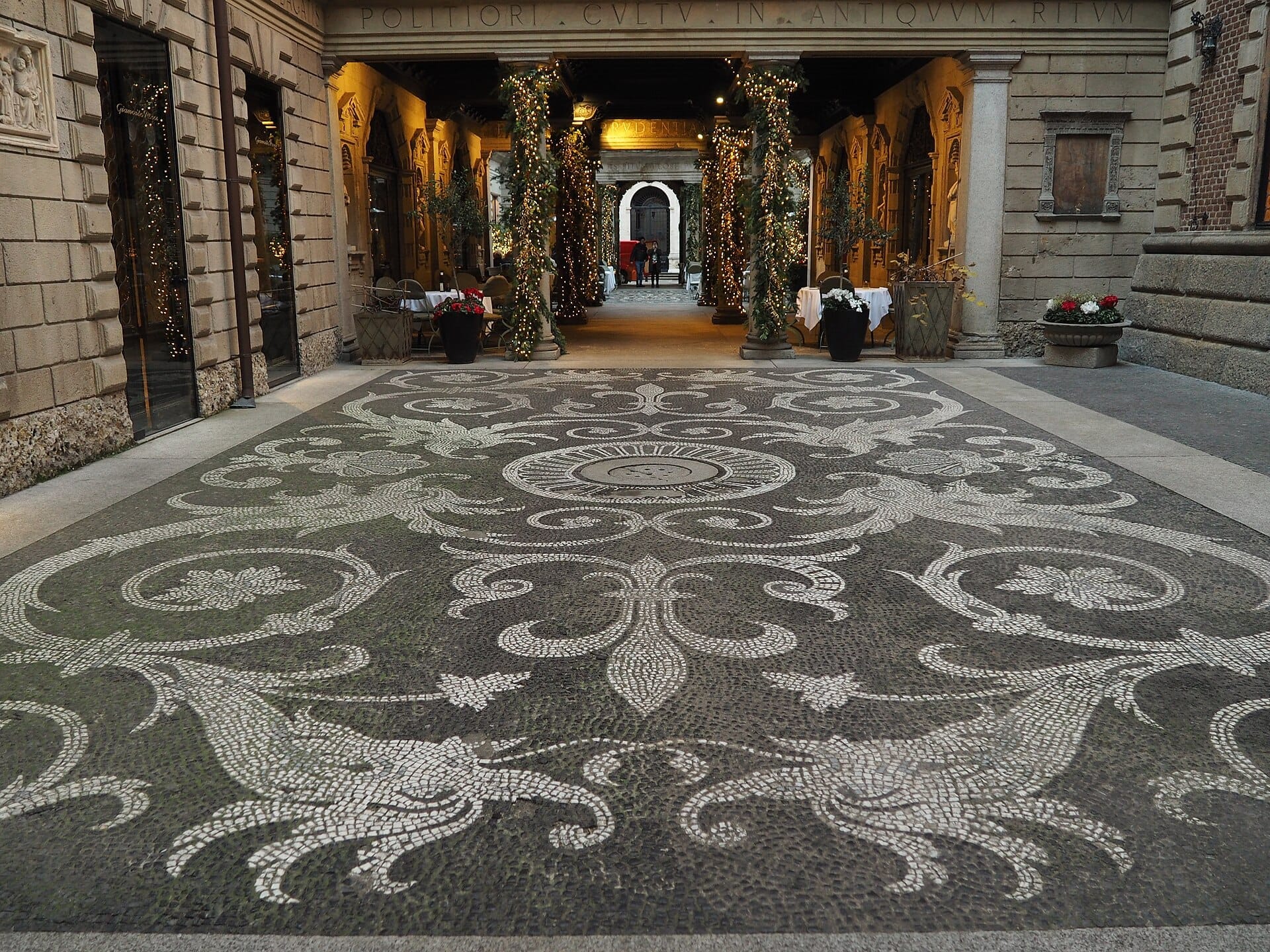 Elegante ingresso con cortile della Casa Museo Bagatti Valsecchi a Milano con intricato pavimento a mosaico e archi illuminati, circondati da storici edifici in pietra.