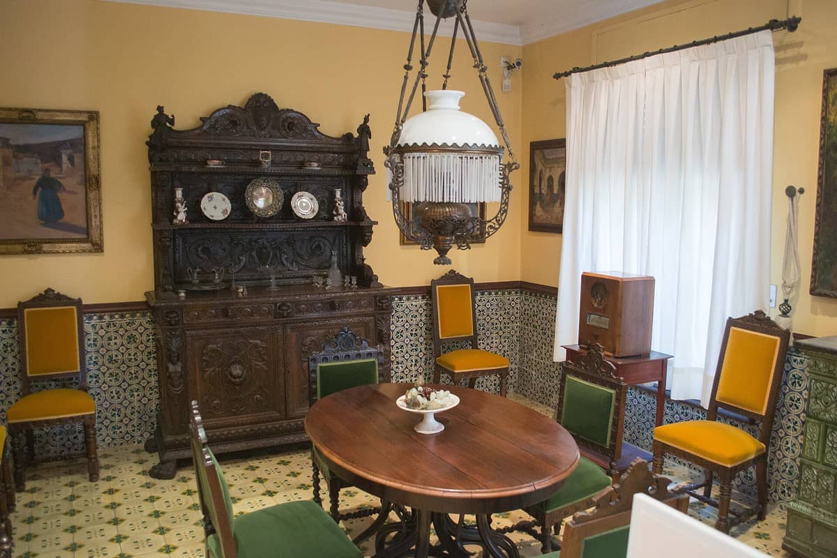 Sala da pranzo della Casa-Museo Benlliure con mobili in legno ornati, pareti di piastrelle colorate, sedie vintage, dipinti e luce naturale attraverso una finestra con tende.
