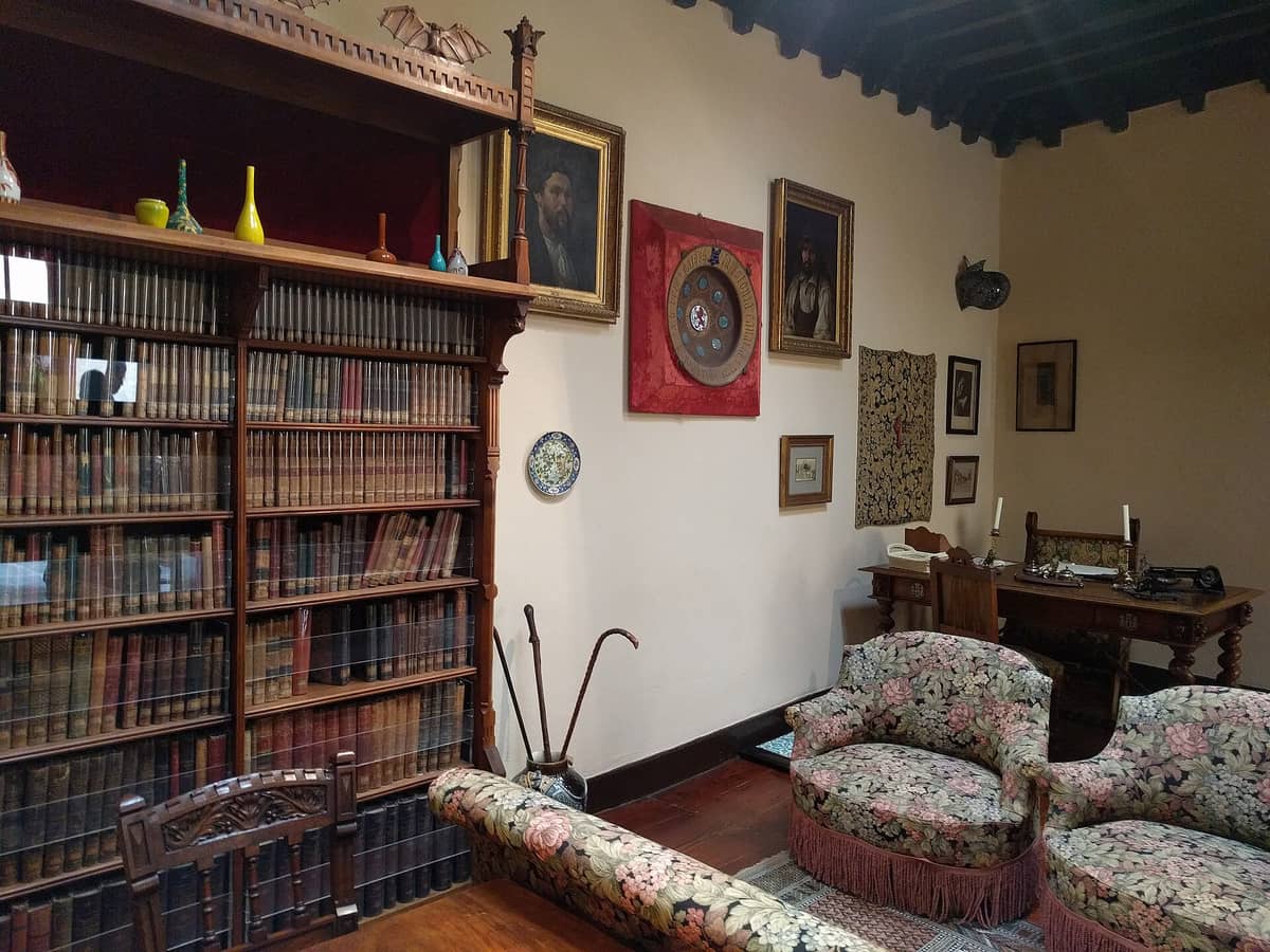 Уютная комната эпохи в Casa-Museo Pérez Galdós: кресла в цветочный принт, большой книжный шкаф со старыми книгами, портреты в рамах и антикварный декор.