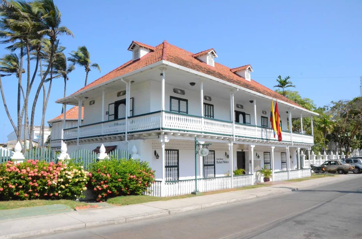 Casa Museo Rafael Núñez with its white wooden facade, red-tiled roof, and Colombian flags, set beside palm trees on a sunny day.