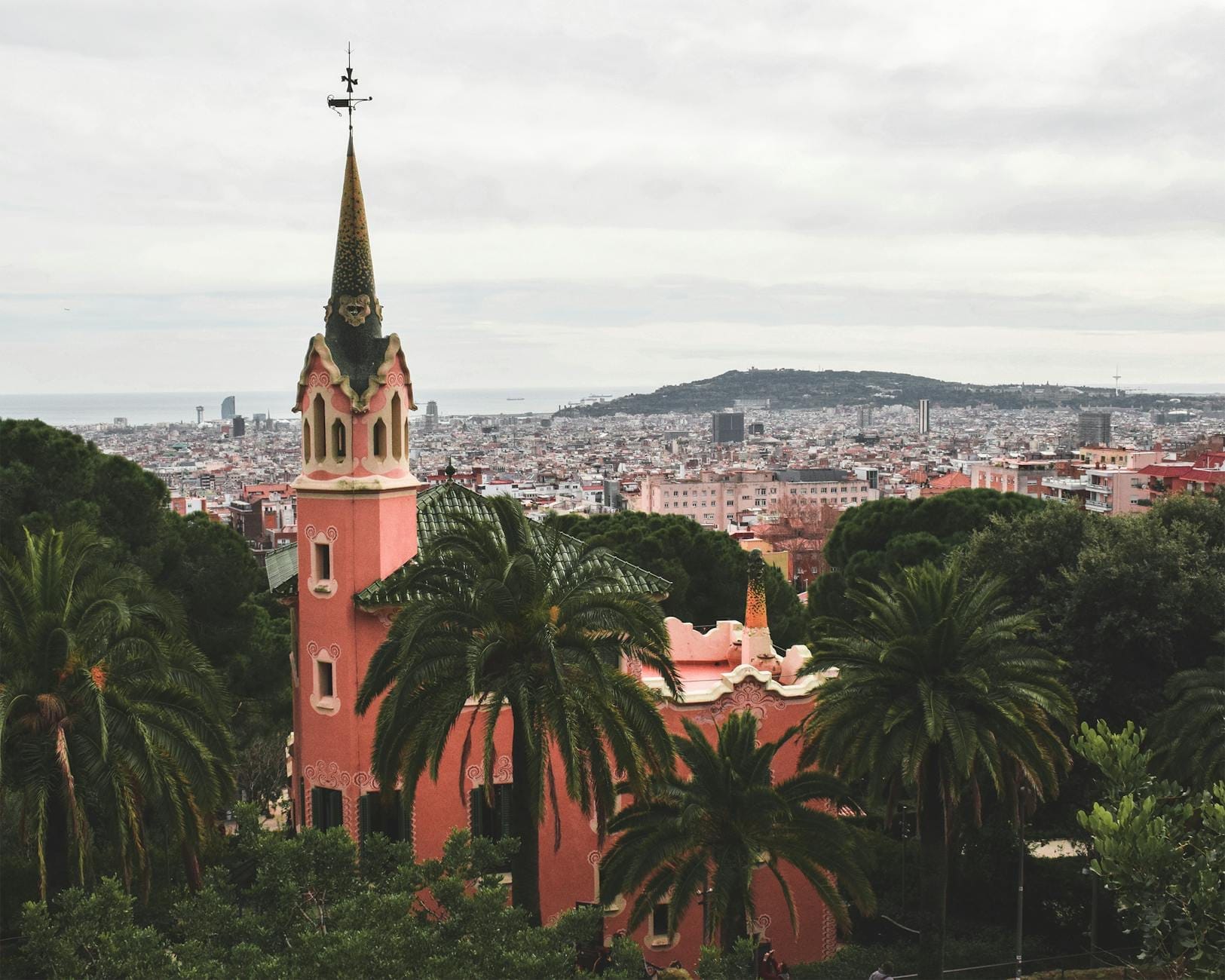 Casa-Museu Gaudí, a pink Neo-Gothic house with a tall spire, surrounded by lush greenery and overlooking the city of Barcelona.
