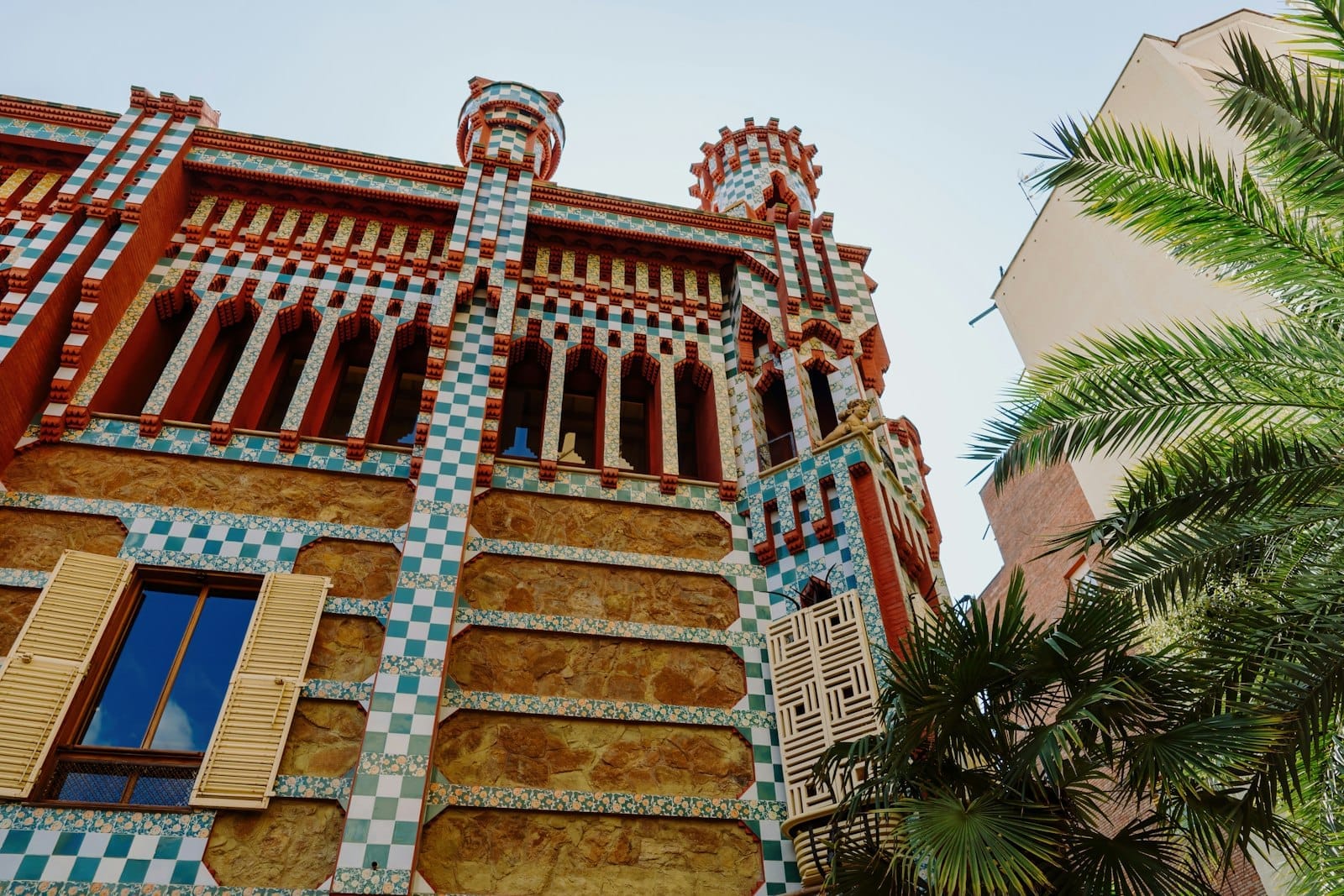 Facade of Casa Vicens with colorful ceramic tiles, intricate brickwork, arched windows, and palm trees against a clear blue sky.