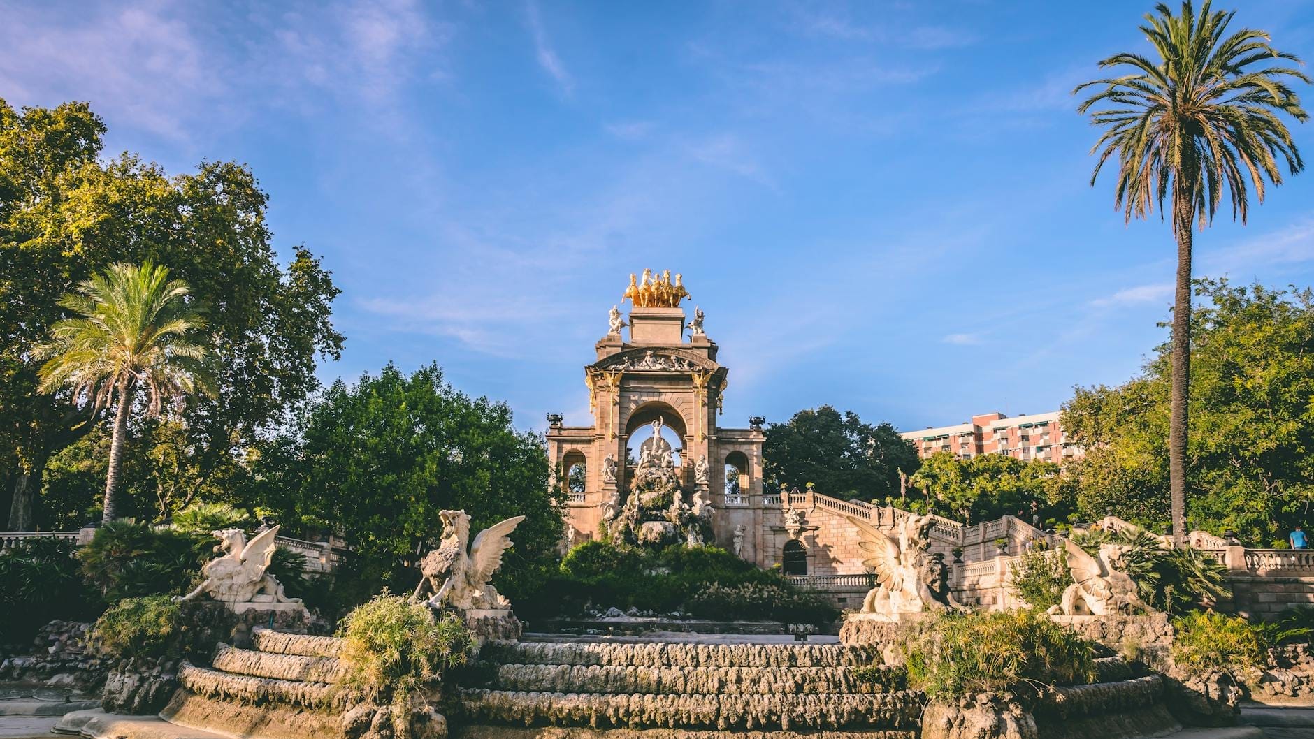 Wide view of Cascada Monumental fountain in Parc de la Ciutadella, Barcelona, with lush greenery, ornate sculptures, turquoise water, and a golden statue under a blue sky.