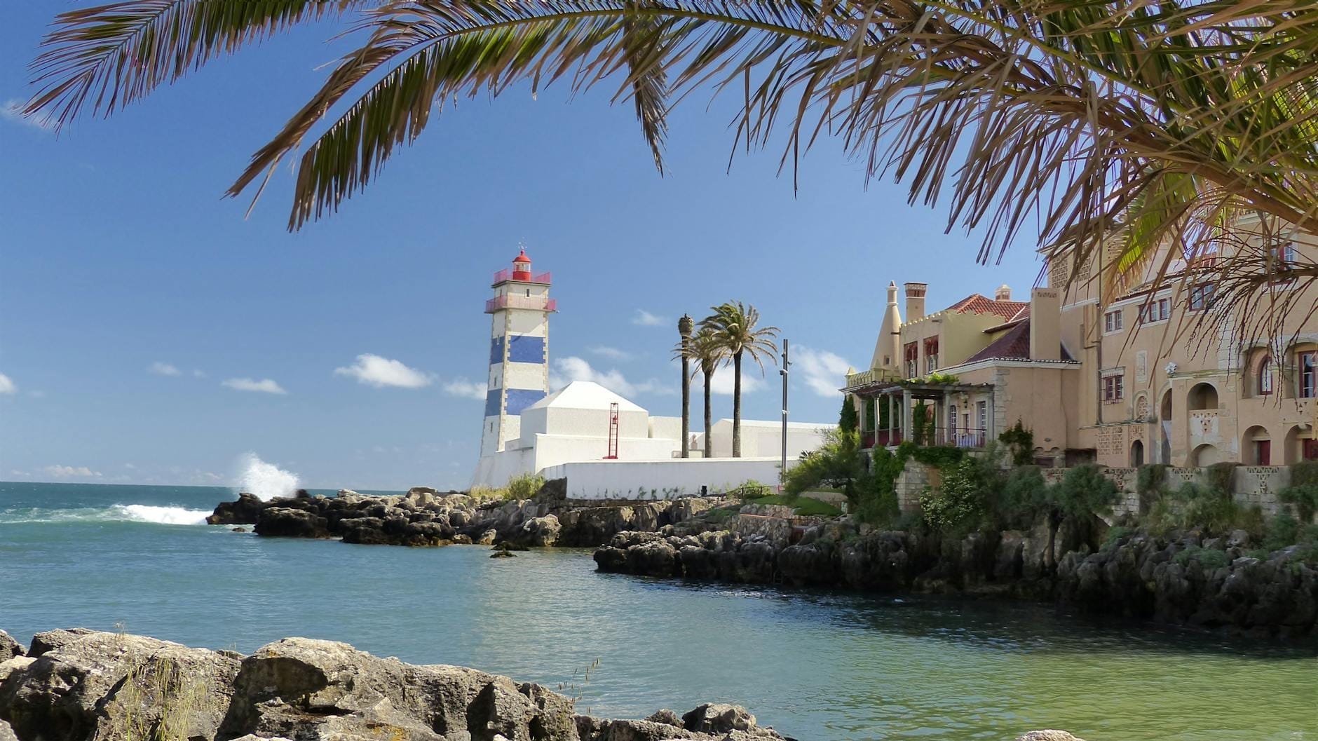 Vista sulla costa di Cascais con il caratteristico faro di Santa Marta, eleganti ville sul lungomare, palme e le acque turchesi dell'Atlantico in una giornata di sole.