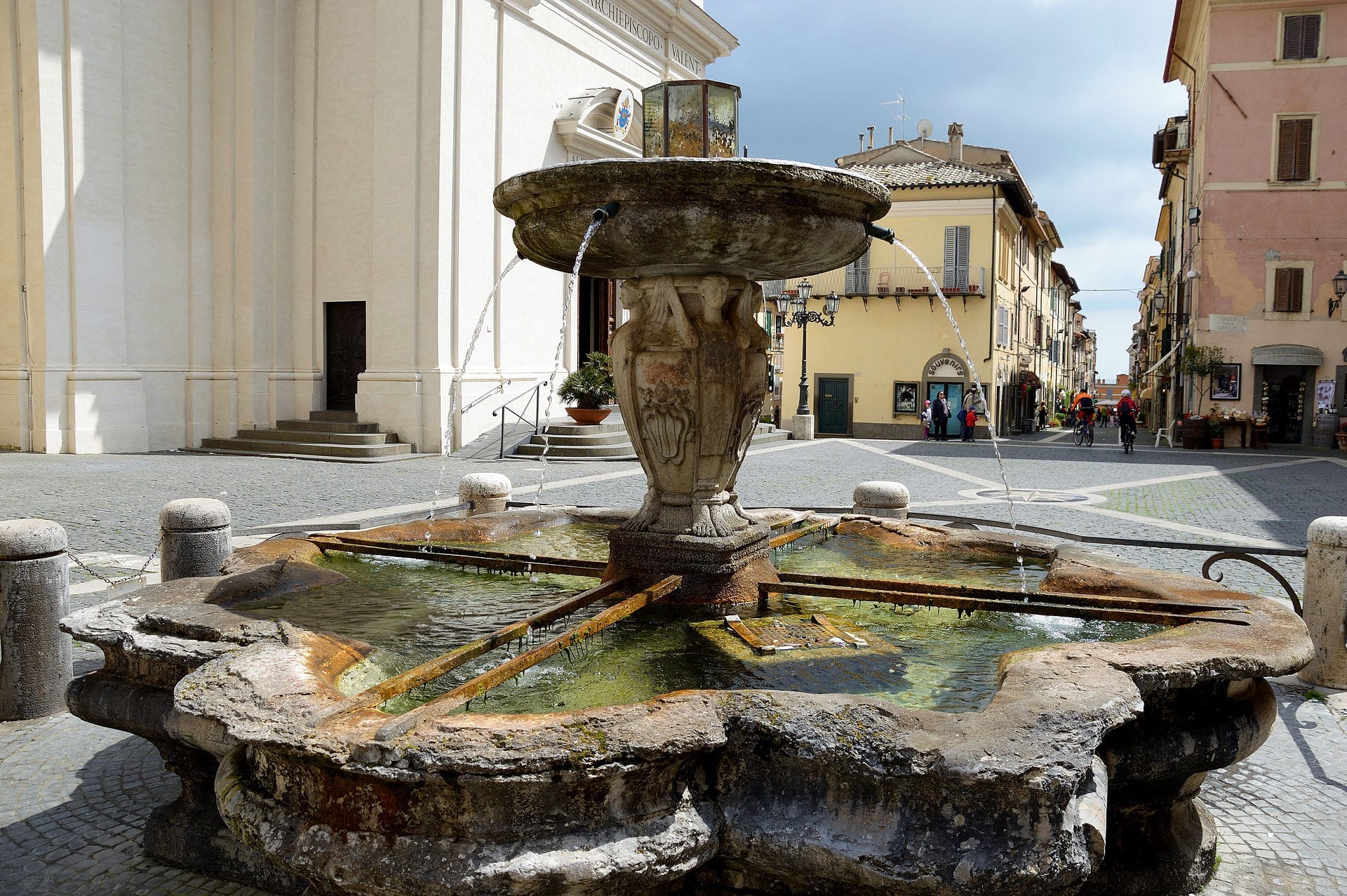 Historic stone fountain in the main square of Castel Gandolfo, with water streaming and classic Italian buildings in the background.