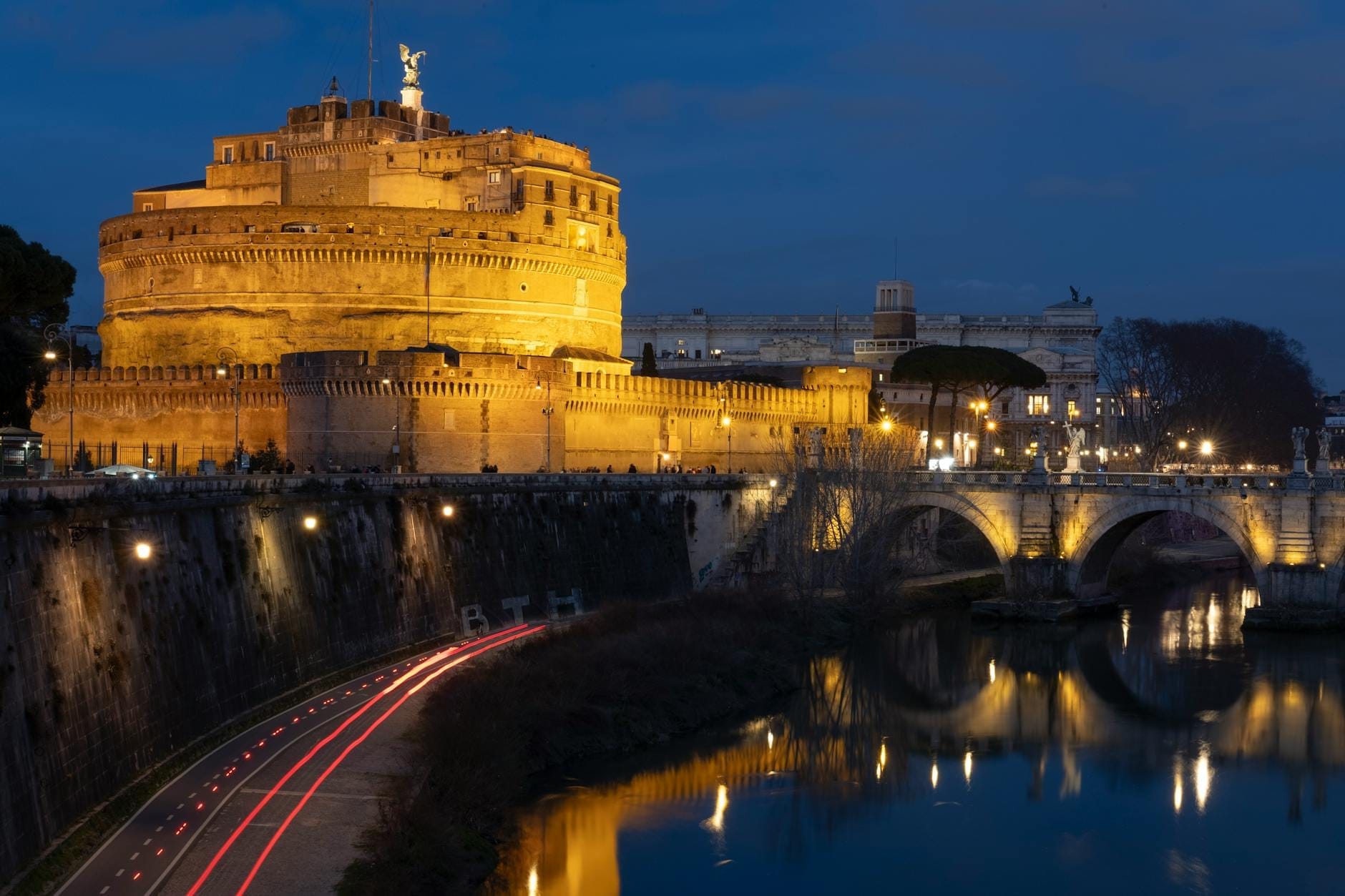Castel Sant'Angelo dramatically illuminated at night, reflected in the Tiber River with the bridge and city lights creating a magical twilight atmosphere.