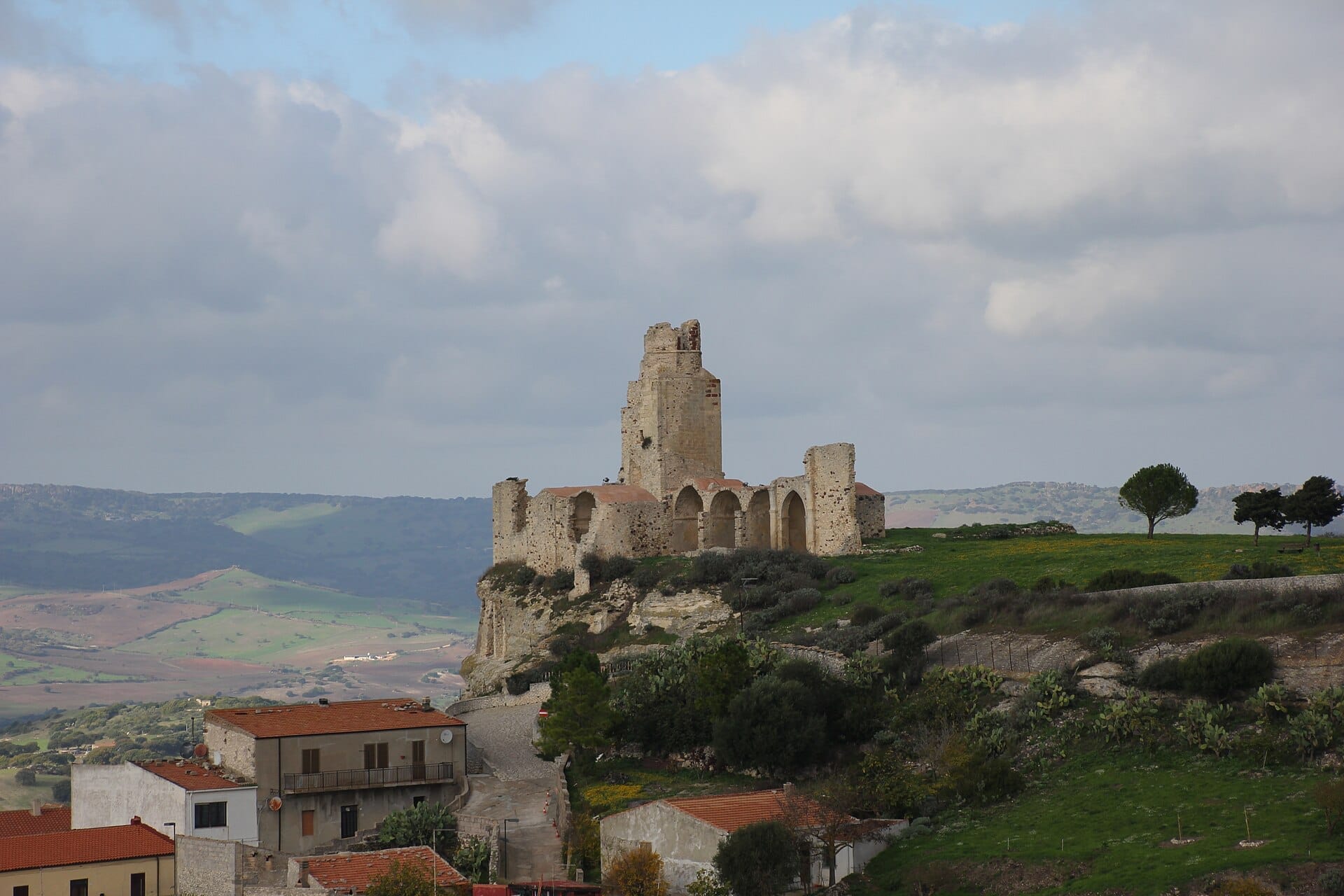 Die Steinruinen des Castello dei Doria erheben sich auf einem grünen Hügel mit Blick auf Häuser und sanfte Hügel unter einem bewölkten Himmel in Castelsardo.