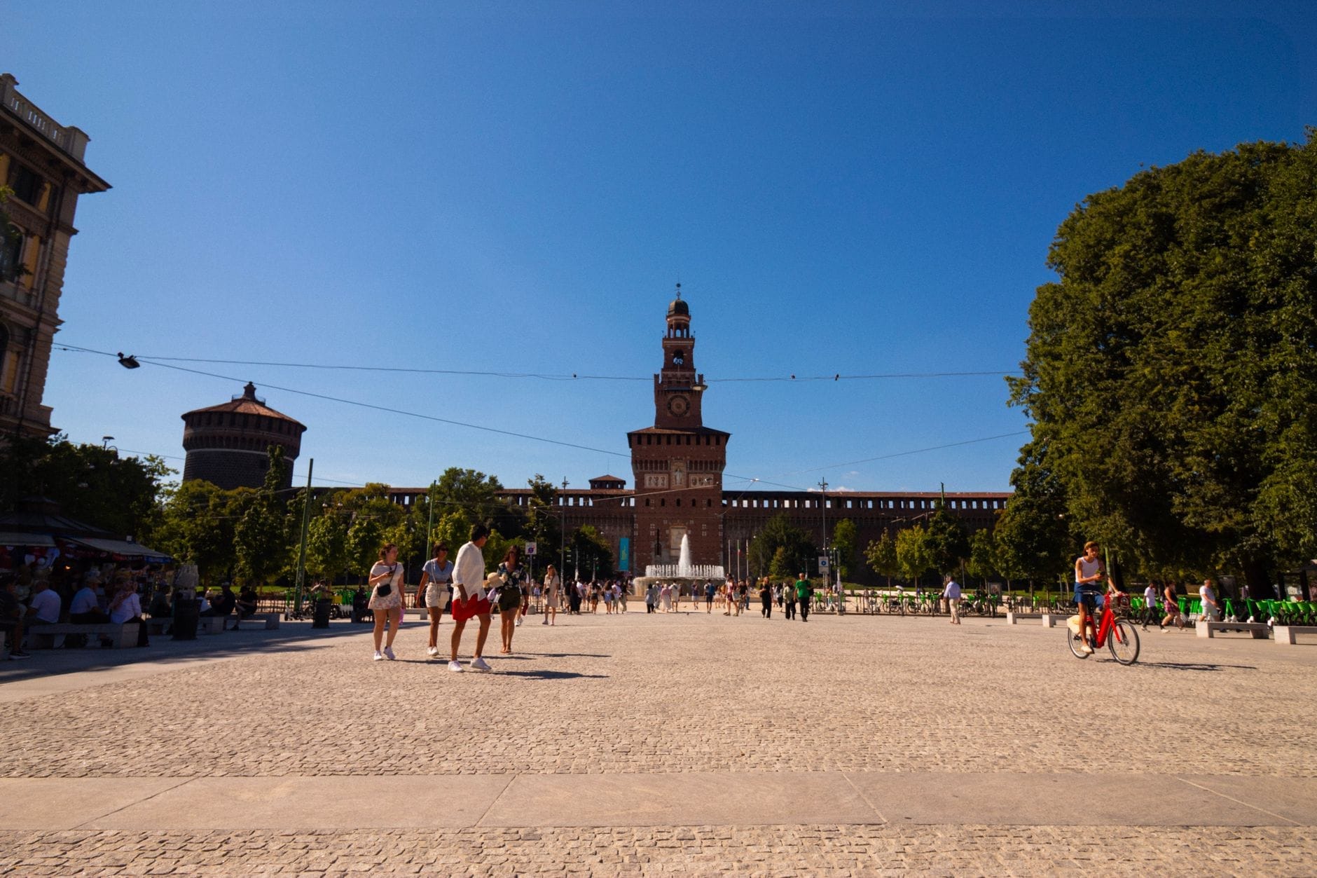 Vista ampia della facciata principale del Castello Sforzesco in una giornata di sole, con visitatori a piedi e in bicicletta sulla grande piazza acciottolata, incorniciata da alberi rigogliosi.
