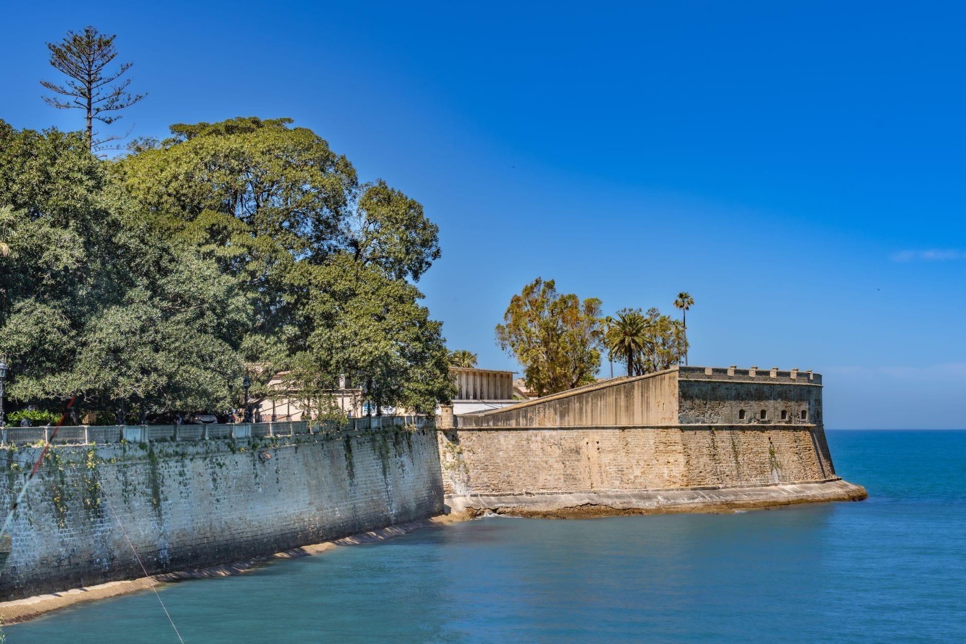 Stone ramparts of Castillo de Santa Catalina extend over blue sea on a sunny day, with lush trees and clear sky in Cádiz, Spain.