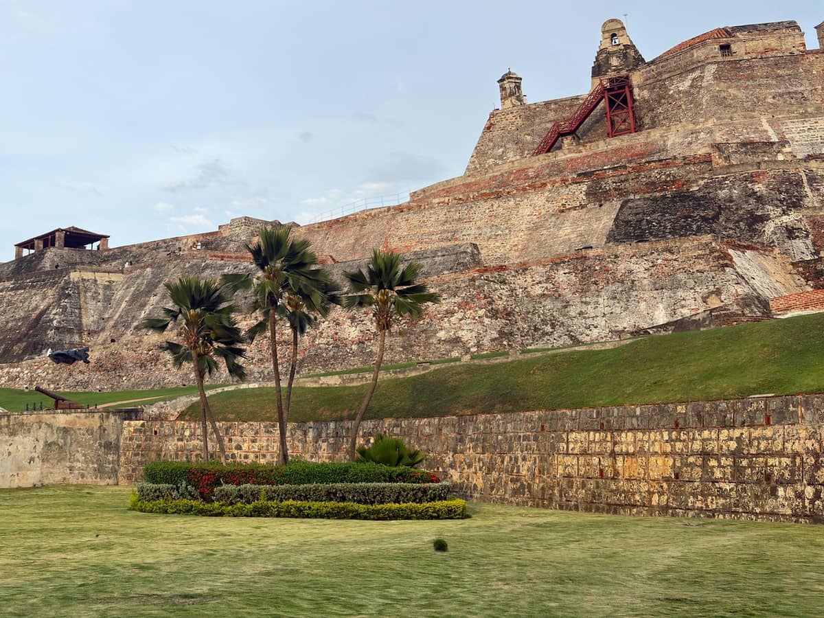Wide view of Castillo de San Felipe de Barajas with weathered stone ramparts, lush green lawn, palm trees, and partly cloudy sky in Cartagena.