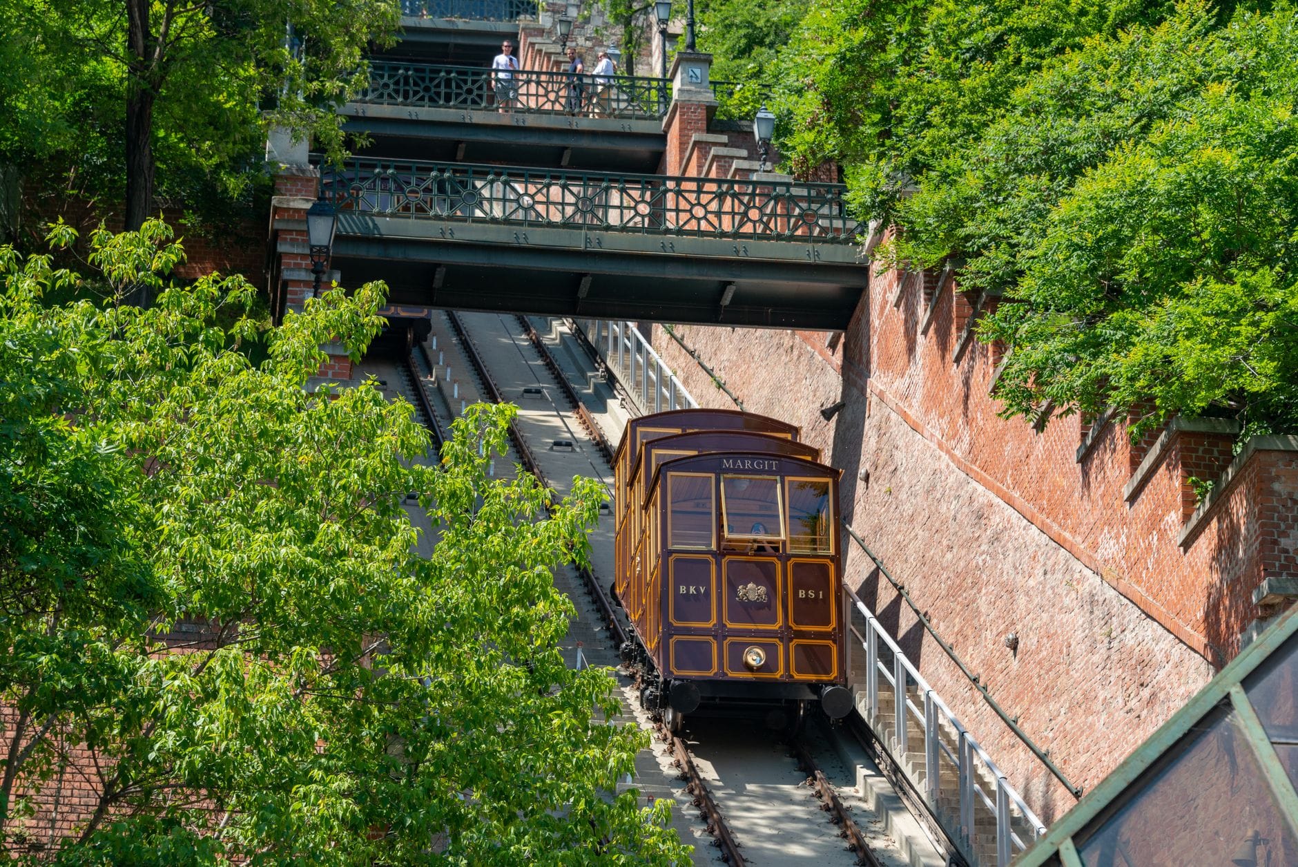 La storica cabina marrone del Budavári Sikló che sale sui binari fiancheggiati da muri di mattoni e alberi rigogliosi sul Colle del Castello a Budapest.