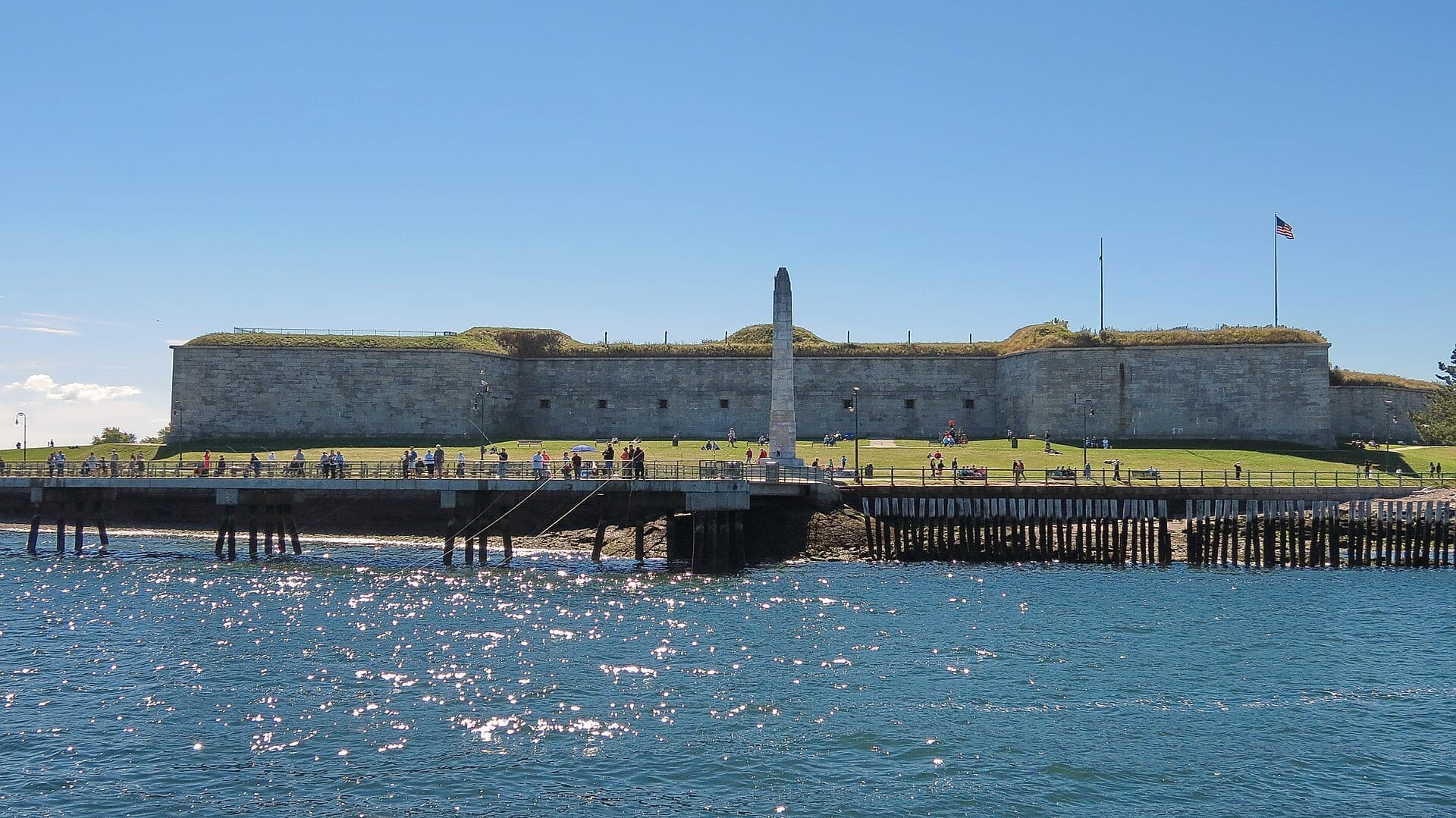 Fort Independence si trova su Castle Island con mura in pietra illuminate dal sole, un prato verde, un monumento a obelisco e visitatori che godono del lungomare sotto cieli azzurri.