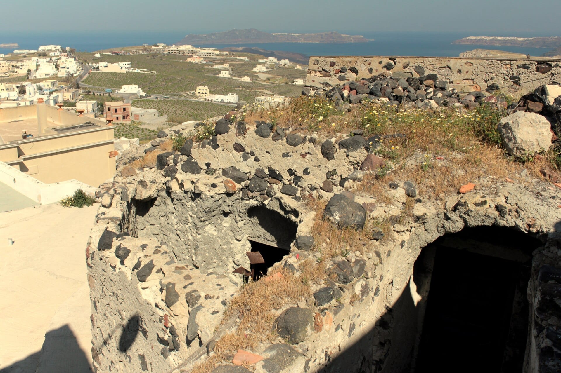 ซากปรักหักพังหินของปราสาท Pyrgos ที่มีดอกไม้ป่า มองเห็นบ้านทาสีขาว ทุ่งหญ้าสีเขียว และทะเลอีเจียนสีฟ้าใน Santorini