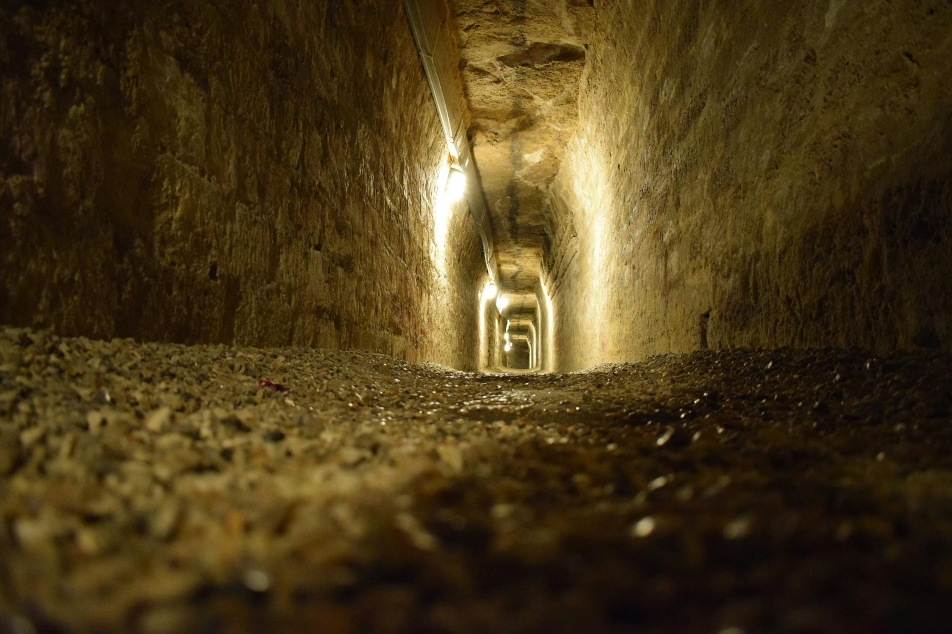 View down a dimly lit, ancient stone tunnel underground with rough gravel floor and walls, evoking the historic passageways of the Catacombs of San Callisto in Rome.