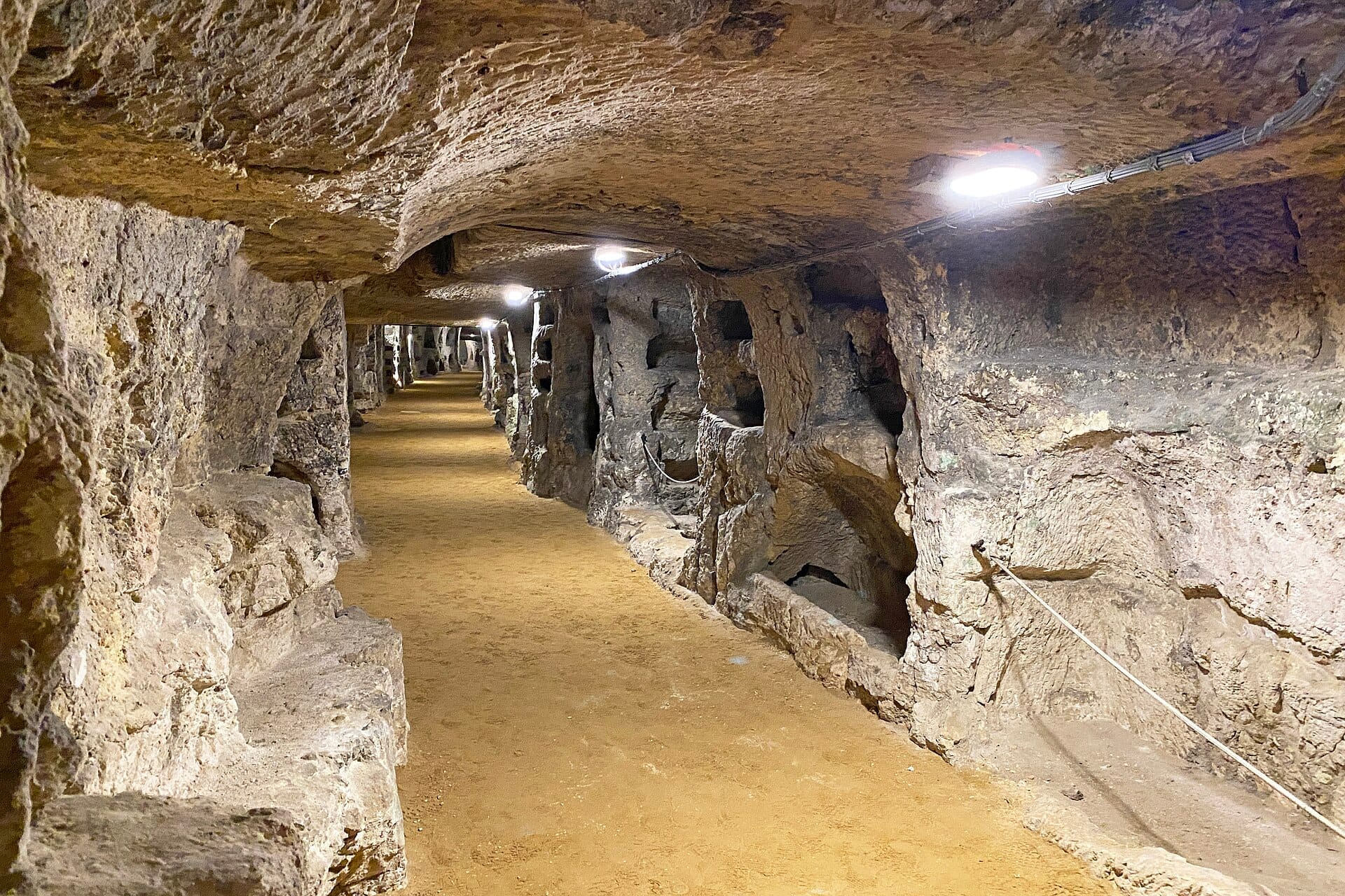 In den Fels gehauener unterirdischer Korridor in den Katakomben von San Giovanni, mit beleuchteten Wänden, Nischen und einem sandbedeckten Gehweg.