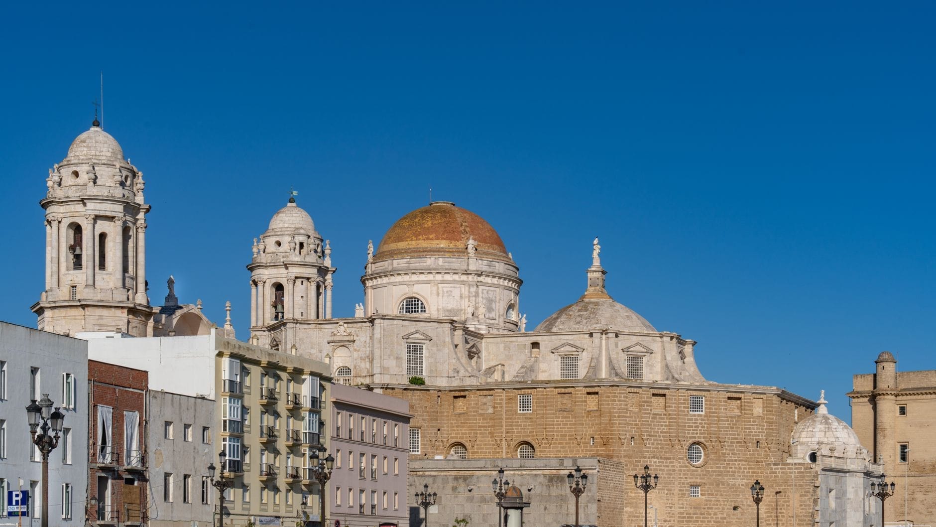 Panoramic view of the Catedral de Cádiz with its golden dome and towers under a clear blue sky, framed by nearby historic buildings.