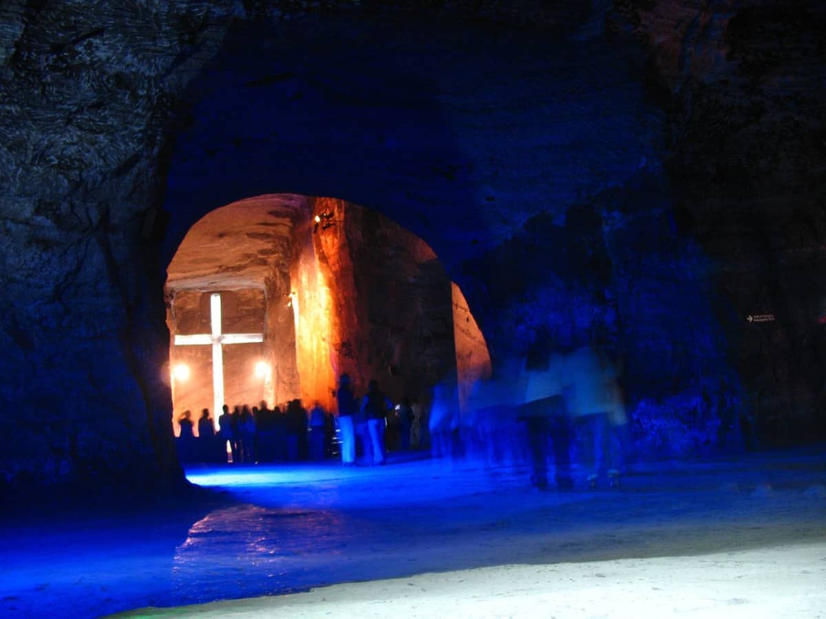 A view inside the Catedral de Sal de Zipaquirá shows a large illuminated cross carved into salt rock and visitors beneath blue lighting.
