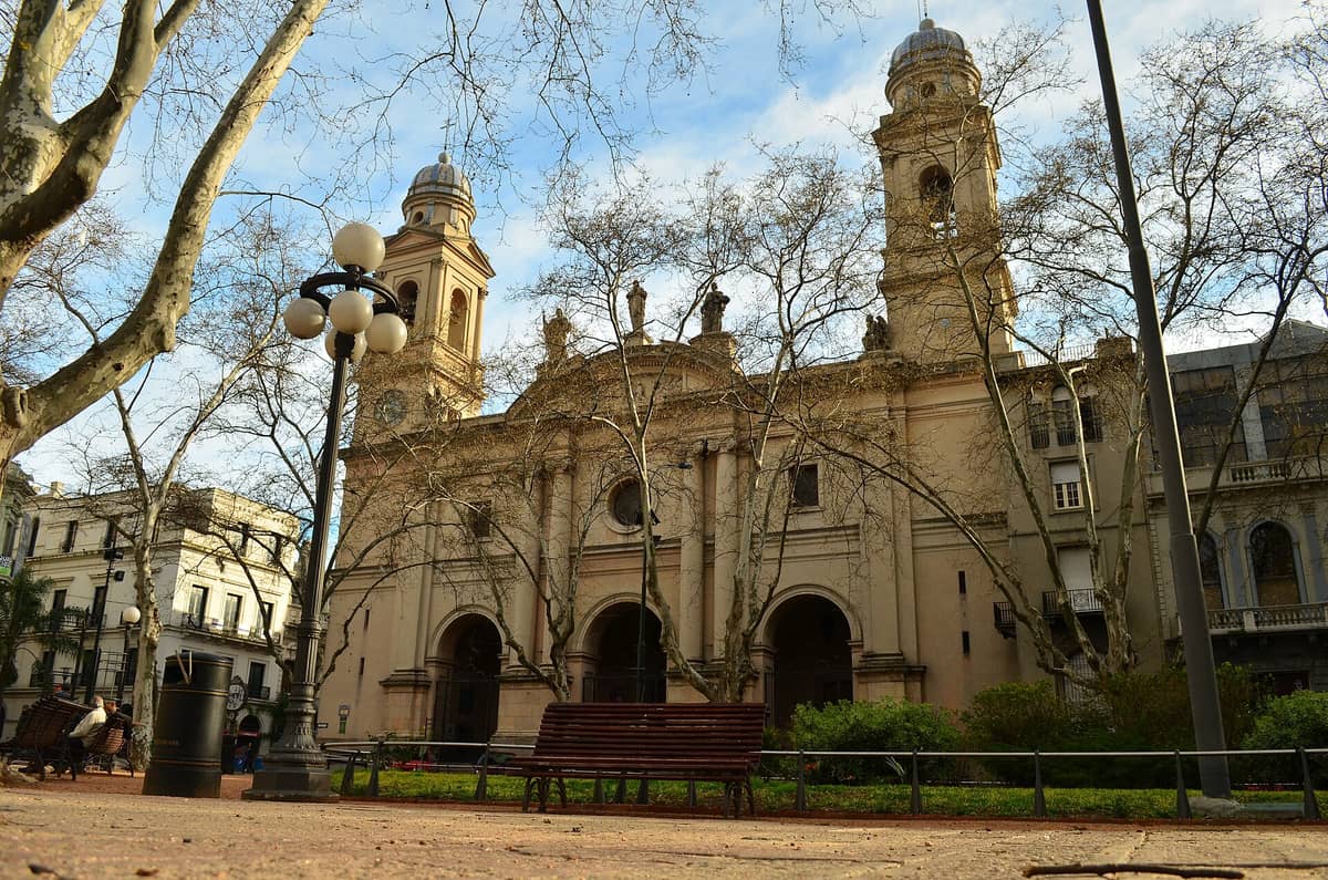 มุมกว้างของด้านหน้าโบสถ์ Catedral Metropolitana de Montevideo สไตล์นีโอคลาสสิกพร้อมหอคู่ ตัดกับต้นไม้ใน Plaza de la Constitución ย่าน Ciudad Vieja