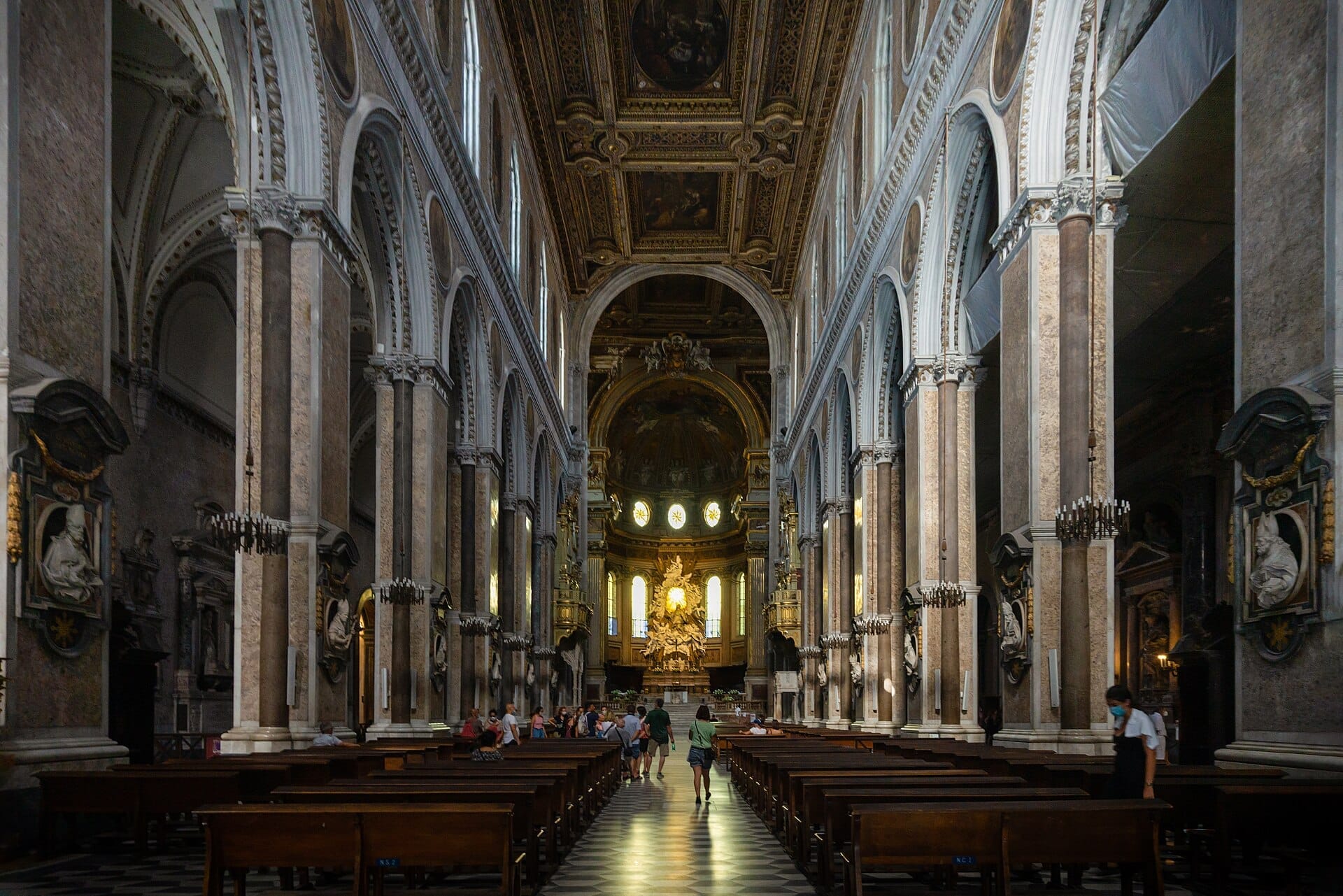 Navata centrale del Duomo di Napoli con alte colonne di marmo, archi ornamentali e visitatori che ammirano l'altare illuminato nella morbida luce del giorno.