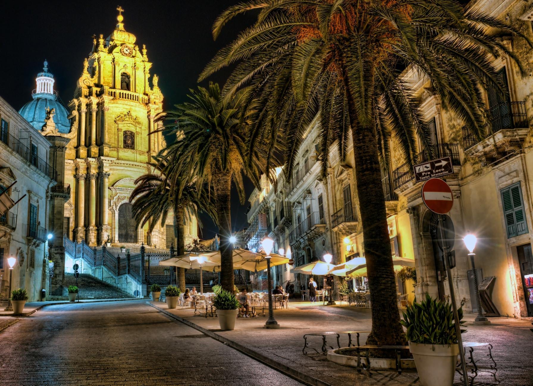 Abendansicht der Kathedrale San Giorgio in Ragusa Ibla, dramatisch beleuchtet mit ihrer Barockfassade und Kuppel, die sich über die palmengesäumte Piazza Duomo und die umliegenden Cafés erhebt.