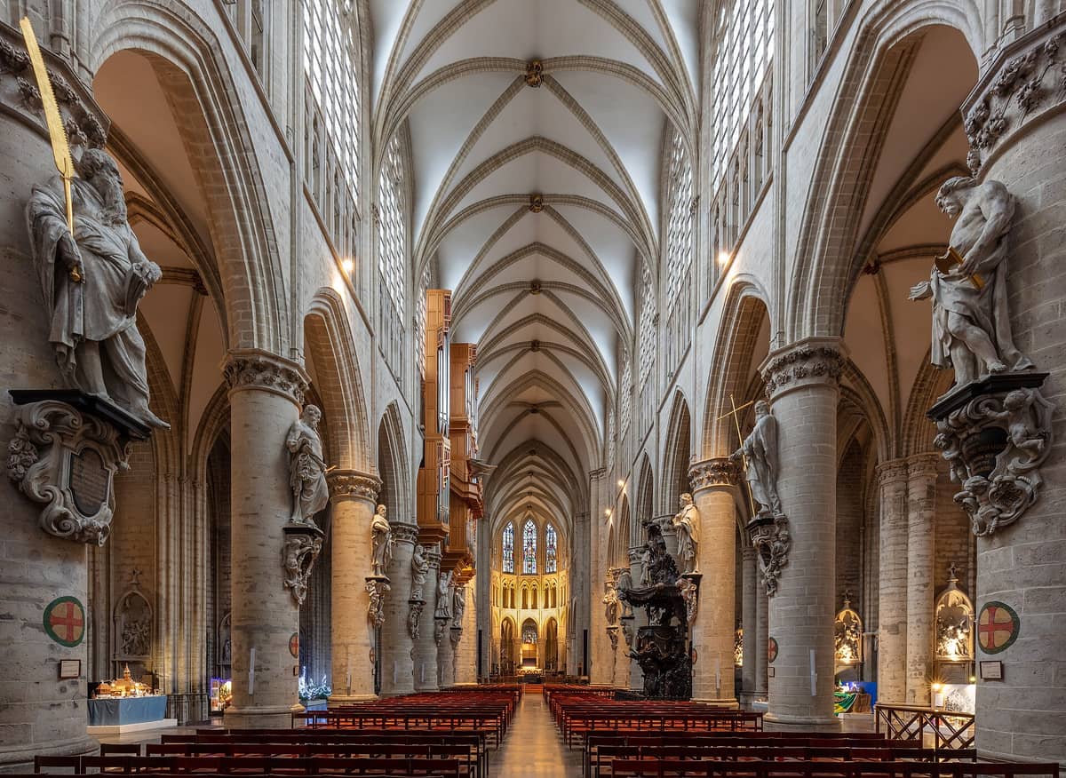 A nave da Catedral de São Miguel e Santa Gudula em Bruxelas, com arcos góticos imponentes, estátuas, vitrais coloridos e fileiras de bancos.